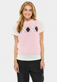 Débardeur en tricot rose sans manches avec un motif argyle dans les tons marron, blanc et bleu clair. Porté par-dessus une chemise à manches courtes crème en tissu texturé.