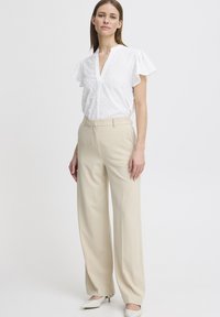 Blusa bianca con decorazioni testurizzate, scollo a V e maniche fruscianti; pantaloni beige a vita alta; scarpe bianche a punta; sfondo neutro.