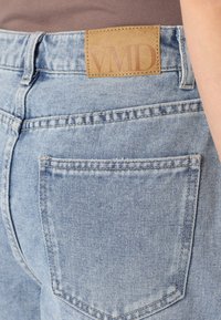 Jeans de denim de color azul claro con un acabado texturizado, que presentan un bolsillo trasero visible y una etiqueta de cuero marrón marcada "VMID" en la cinturilla.