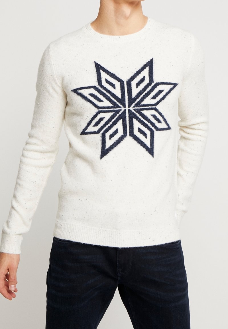 Suéter blanco de punto con un gran diseño geométrico de copo de nieve en color azul marino, que presenta un cuello redondo y puños y dobladillo acanalados.