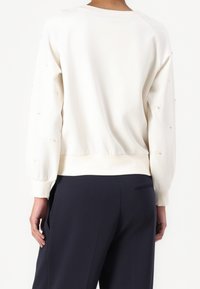 Persoon die een wit shirt met lange mouwen draagt met subtiele borduursels op de mouwen en donkerblauwe broek, gezien van achteren.