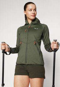 Olivegrønn jakke med glidelås og hette, med oransje glidelåstrekk og sidelommer. Brukes med matchende shorts, holder på trekkingstaver.