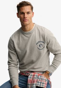 Szara bluza z okrągłym dekoltem z niebieskim nadrukiem z napisem "MERCHANT No. 1 TRADING CO." Materiał o fakturze z prążkowanymi mankietami. Noszona z koszulą w kratę.