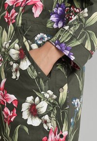 Main reposant à l'intérieur d'une poche de veste florale verte ornée de fleurs blanches, rouges, roses, violettes et bleues avec des feuilles vertes.