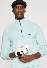 Camisola com fecho de correr azul-claro com detalhes a preto, apresentando um padrão texturizado, logótipo no peito e uma luva de golfe na mão.