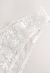 Lingerie en dentelle blanche avec broderies florales, tissu en maille transparente, bords festonnés et galon décoratif picot le long de la ceinture.