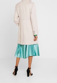 Donna che indossa un cappotto beige fino al ginocchio sopra una gonna di raso teal e scarpe con tacco alto nere con cinturino alla caviglia, in piedi di spalle.