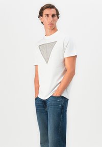 Jeune homme aux cheveux bouclés portant un t-shirt blanc avec le logo triangle GUESS et un jean bleu, les mains dans les poches, sur un fond blanc uni.