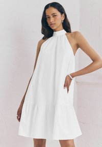 Abito bianco a collare halter realizzato in tessuto leggero, con scollatura arricciata e orlo a balze a strati. Due tasche laterali visibili.