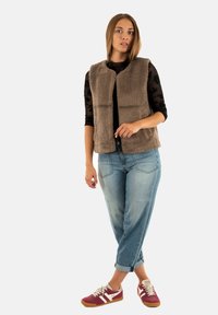 Una giovane donna indossa un gilet di pile marrone sopra una maglietta nera trasparente, jeans azzurri arrotolati e sneakers rosse con strisce bianche.