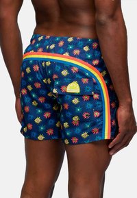 Una persona con un fisico muscoloso indossa un costume da bagno colorato con un motivo a sole, con una striscia arcobaleno lungo il lato.