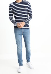 Marinblå långärmad randig t-shirt med vita ränder, rund hals, liten logotyp på bröstet; kombinerad med ljusblå jeans och vita sneakers.