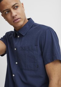 Camisa de botones de manga corta en tejido texturizado de color marino. Presenta un bolsillo en el pecho y botones blancos. Corte entallado con cuello.