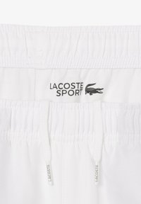 Weiße Shorts mit elastischem Bund, ausgestattet mit einem schwarzen "LACOSTE SPORT" Logo und strukturiertem Stoff. Einschließlich Kordeln zur Anpassung.