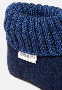 Pantofola di feltro blu scuro con un polsino a costine in tonalità di blu. Presenta un'etichetta con la scritta "Gottstein." Texture morbida e design informale.