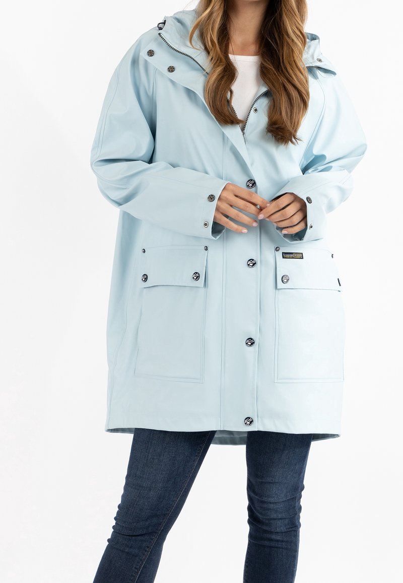 Imperméable bleu clair à capuche avec boutons devant et grandes poches à rabat, porté sur un jean skinny foncé.