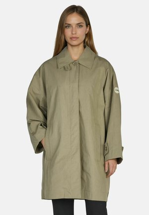 Woman wearing oversized light olive green coat with collar and buttoned cuffs, hands in pockets, facing forward on plain background.