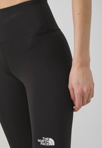 Svarta sportleggings med hög midja, med en vit logotyp på vänster sida och en slät yta. Inga synliga sömmar eller mönster.