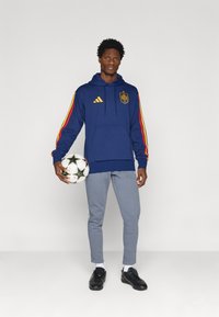 Ναυτικό φούτερ με κουκούλα και κόκκινες-κίτρινες μανίκες, που διαθέτει λογότυπο και τσέπη καγκουρό, συνδυασμένο με ανοιχτό γκρίζα παντελόνια και μαύρα παπούτσια.