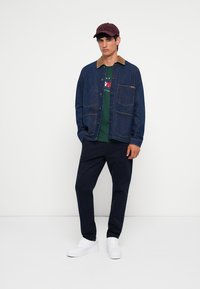 Chaqueta de mezclilla con cuello marrón, camiseta gráfica verde, pantalones azul marino y zapatillas blancas. Modelo con una gorra burdeos, de pie frente a un fondo liso.