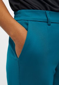 Personne portant un pantalon turquoise à taille élastique avec la main dans la poche latérale, associé à un haut foncé.