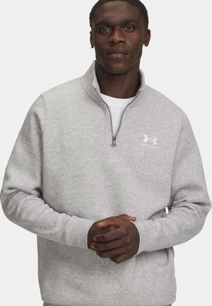 Mann trägt ein hellgraues Under Armour Sweatshirt mit Viertel-Reißverschluss, die Hände verschränkt, blickt nach vorne vor einem schlichten hellen Hintergrund.