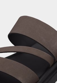 Gros plan d'une sandale noire avec trois larges brides en suède marron traversant le pied.