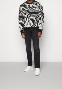 Homme portant un pull à motif abstrait noir et blanc, un jean noir déchiré et des baskets blanches, debout sur un sol gris.