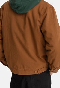 Ruskea kangastakki rentolla istuvuudella, jossa on vihreä huppu ja takana kaksinappinen suljenta. Kankaan pinta on teksturoitu ja saumat näkyvät.