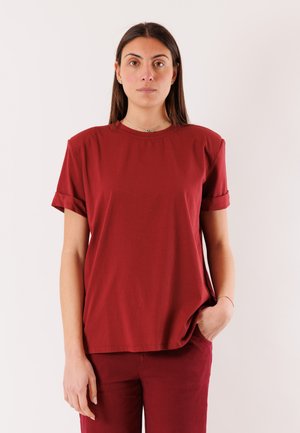 Donna con capelli castani lisci che indossa una maglietta rossa a maniche corte e pantaloni rossi, in piedi con una mano in tasca contro uno sfondo bianco.