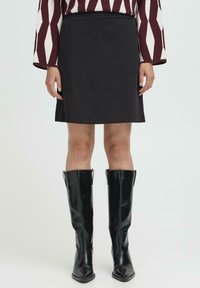 Mujer que lleva una falda negra a la altura de la rodilla, botas negras de caña alta y ajustadas, y una blusa de manga larga con un patrón geométrico en rojo y blanco.