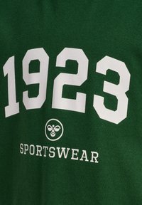 Mørkegrønt stof med store hvide "1923" tal, et bi-logo i en cirkel, og ordet "SPORTSWEAR" trykt nedenfor.