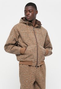 Bruine zip-up hoodie met een cirkelvormig patroon, gevoerde capuchon en voorzakken. Past bij bijpassende broek voor een complete look.