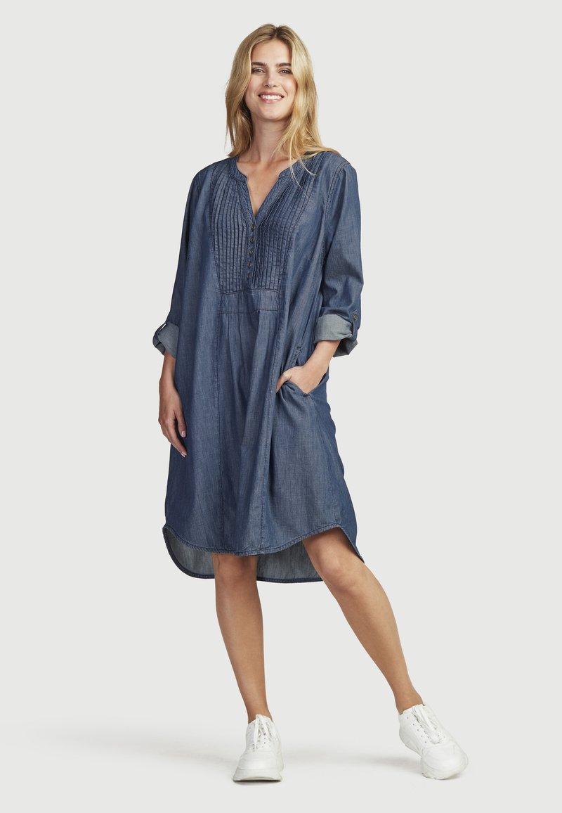 Denim jurk met een V-hals, geplooide voorkant en knoopdetails. Lange mouwen zijn opgerold. Voorzien van zijzakken en een high-low zoom.