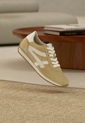 Beige suède sneaker met witte lederen accenten en veters, voorzien van een gestructureerde rubberen zool en een kenmerkend zijontwerp.