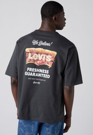 Muž nosi čierne tričko s veľkým obrázkom kúsku pizze a bielym textom: „Dodávame! Čerstvosť Levi's zaručená.”