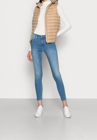 Beżowa kamizelka puchowa z kapturem założona na białą golfową bluzkę, w połączeniu z jasnoniebieskimi obcisłymi dżinsami i białymi trampkami. Prosty i dobrze dopasowany fason.