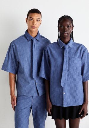 Two people stand side by side wearing matching blue checkered short-sleeve shirts with ties; one wears matching pants, the other a black skirt.