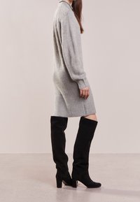 Robe grise en maille côtelée avec manches longues, dotée d'un col montant. Associée à de hautes bottes en daim noir, soulignant la silhouette de la tenue.