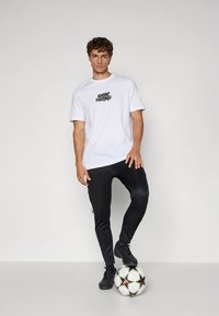 T-shirt en coton blanc avec le texte noir "Adidas Football", assorti à un pantalon noir avec des bandes blanches sur les côtés, debout sur un ballon de football à motif.