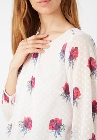 Blusa blanca transparente con lunares, con patrones florales rojos. Escote redondo, mangas largas y botones dorados en el hombro.