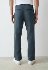 Mann in dunkelgrauen geraden Hosen und weißen Sneakers, der vor einem einfachen hellen Hintergrund steht, von hinten betrachtet.