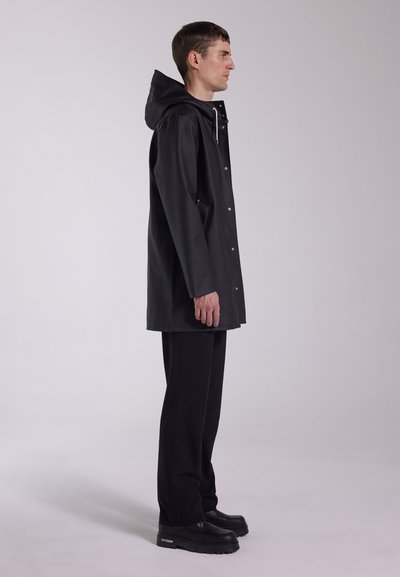 Svart vattentät regnrock av slätt material, med huva, tryckknappar och sidofickor. Bärs över svarta byxor och robusta skor.