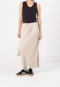 Donna che indossa un top senza maniche nera con bottoni, una gonna maxi beige con cordoncino, sneakers nere, calze bianche e tiene in mano una borsa bianca.
