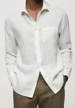 Homme portant une chemise blanche à manches longues avec une poche poitrine et un pantalon marron, mains dans les poches, fond neutre.