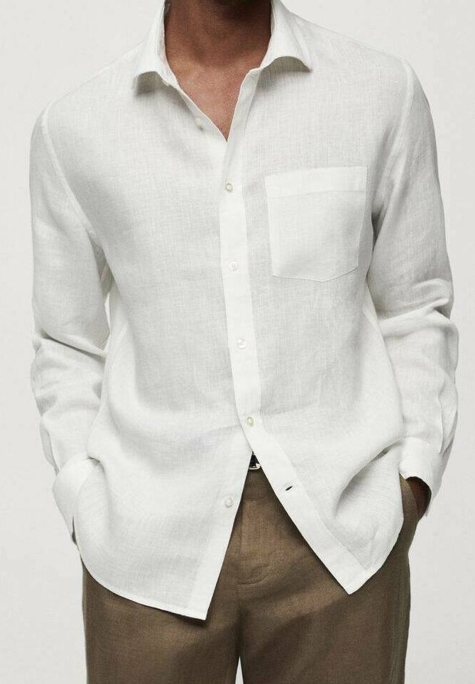 Homme portant une chemise blanche à manches longues avec une poche poitrine et un pantalon marron, mains dans les poches, fond neutre.