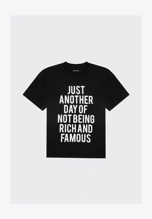 Czarna koszulka z białym napisem "PO PROSTU KOLEJNY DZIEŃ NIE BĘDĄC BOGATYM I SŁYNNYM" dużymi, pogrubionymi literami.