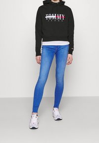 Sudadera negra recortada con el logo "TOMMY JEANS" en blanco, rojo y azul. Combinada con jeans skinny azules y zapatillas blancas. Tejido suave.