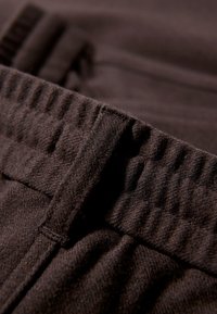 Brązowe materiałowe spodnie z teksturowanym pasem. Pas ma wypukłe prążki, a materiał wydaje się być miękki i elastyczny.