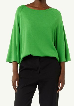 Femme portant un haut ample vert vif à manches larges et un pantalon noir taille haute sur un fond clair.